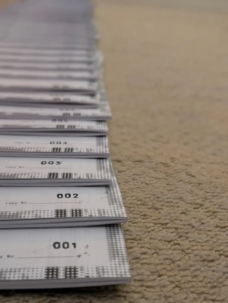 Grey booklets numbered 001 to 004 in a row on a beige carpet, with a shallow depth of field blurring the copies into the distance.