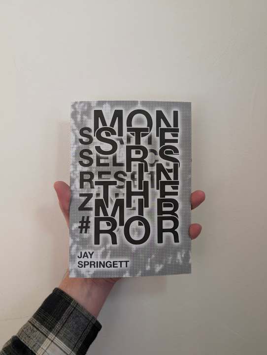 A hand holding the book MONSTERS IN THE MIRROR by Jay Springett, featuring bold black overlapping typography on a halftone cover against a white wall.