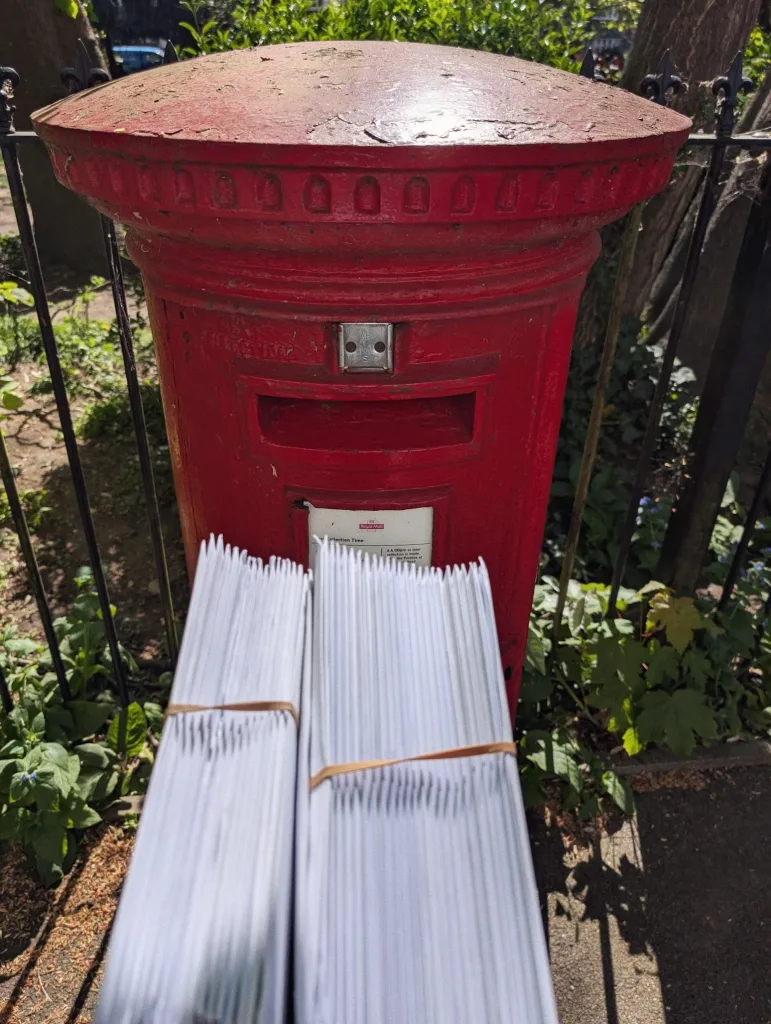 Two thick bundles of white envelopes held by rubber bands in front of a red pillar post box by a park fence.