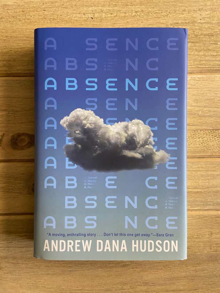 A copy of Absence by Andrew Dana Hudson on a wooden table. The blue cover features the title repeated with missing letters and a small white cloud.