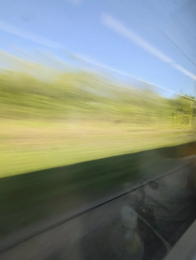 Long exposure horizontal motion blur of green fields and trees rushing past a window under a bright blue sky.