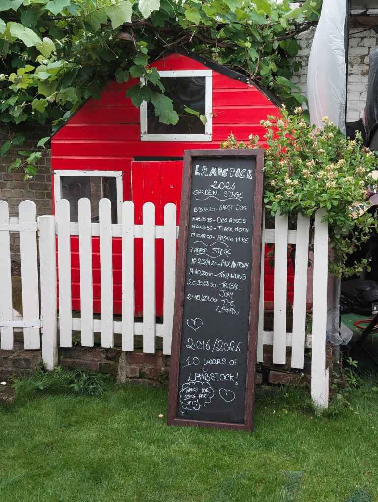 A chalkboard lineup sign for Lambstock 2026 leans against a white picket fence in front of a bright red garden playhouse.
