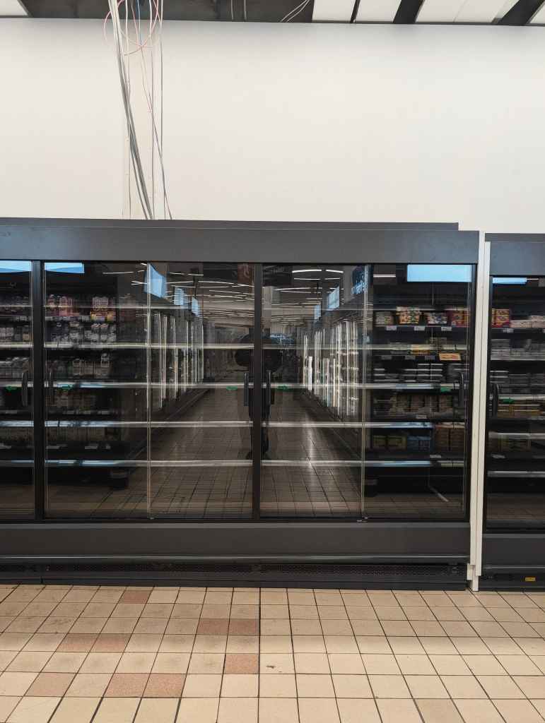 A row of glass-doored supermarket freezers reflecting a long aisle and the silhouette of the person taking the photo.