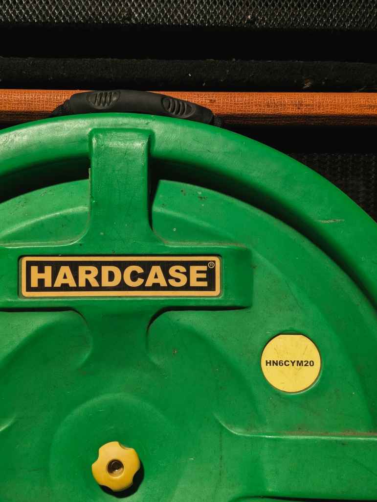 A green Hardcase cymbal case with a yellow logo and a round yellow sticker, set against a black speaker cabinet with an orange stripe.