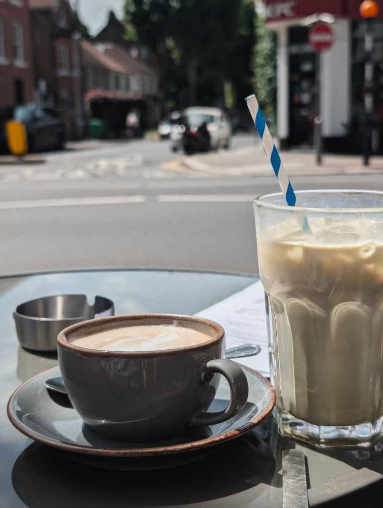 A cappuccino in a grey cup and an iced latte with a striped paper straw on an outdoor cafe table with a blurred street in the background