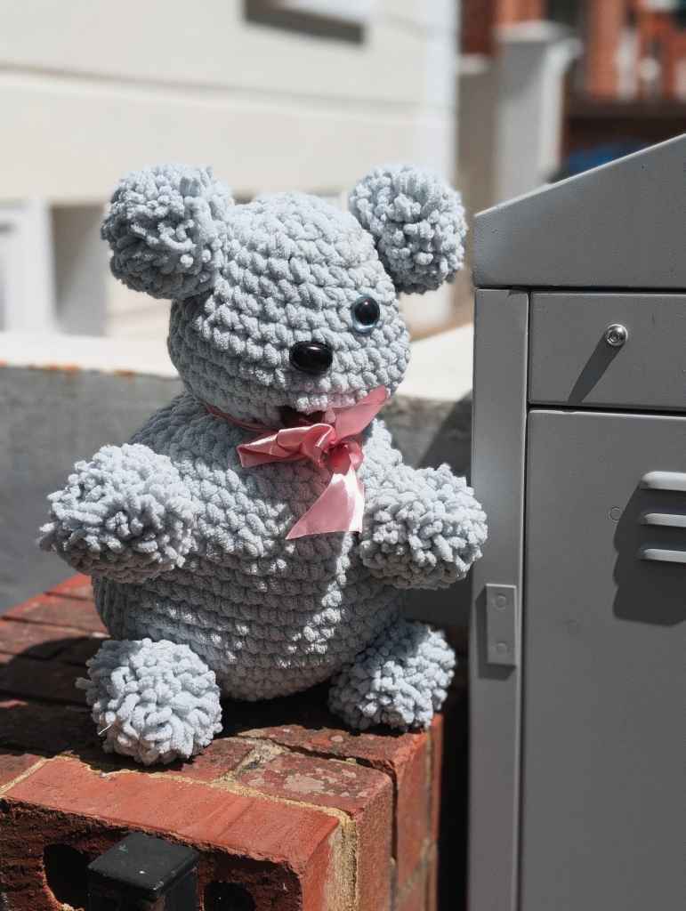 A grey crocheted teddy bear with a pink ribbon sits on a red brick ledge in the sun next to a grey metal cabinet.