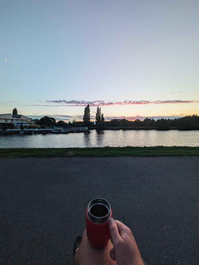 A hand holding a red thermos flask overlooking a wide river at sunset with soft pink and orange light on the horizon