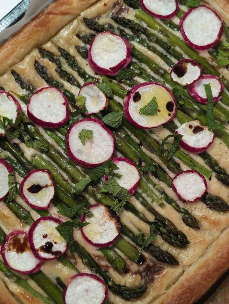 A puff pastry tart topped with neat rows of green asparagus, sliced pink-rimmed radishes, fresh mint, and balsamic glaze.