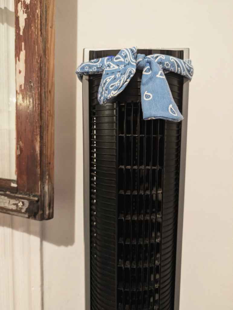 A black tower fan with a light blue paisley bandana tied around its top like a headband, next to a weathered wooden frame