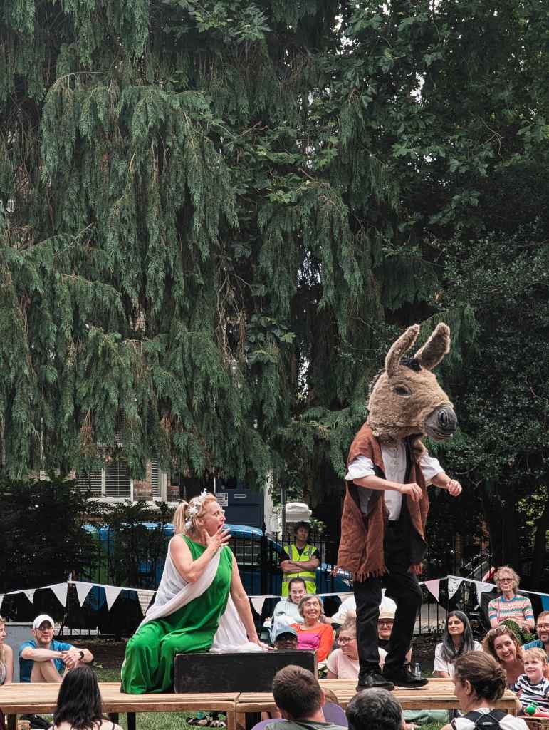 An outdoor theater performance with an actor in a green dress looking startled next to another wearing a large donkey head mask.