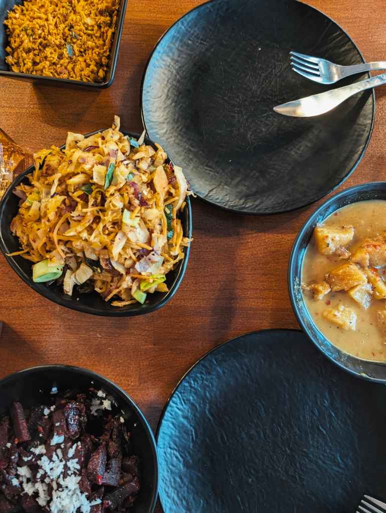Bowls of Sri Lankan kottu roti, beetroot curry, and pol sambol served on a wooden table alongside two empty black textured plates.
