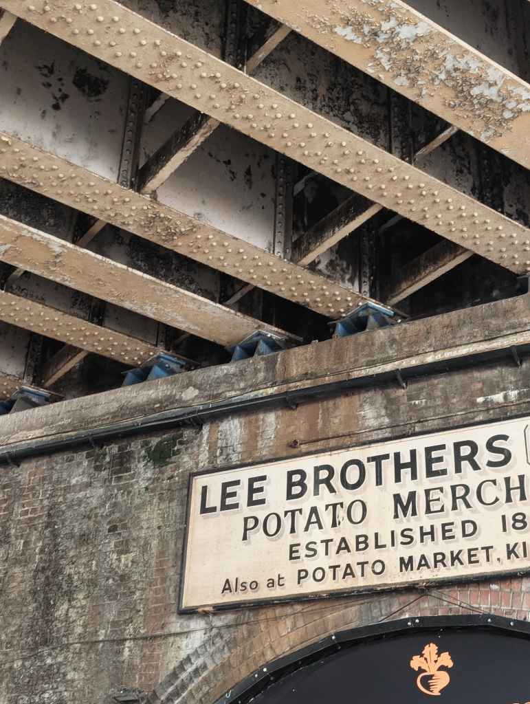 A vintage sign for Lee Brothers Potato Merchants on a brick wall beneath heavy, riveted steel bridge girders.